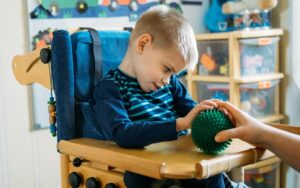 Boy in high chair is playing with sensory ball, carer's hand in shot