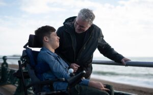 young man in wheelchair at the beach, with an adult, looking happy