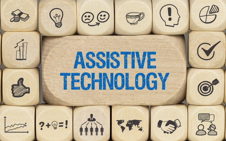 The words 'assistive technology' in blue on wooden rectangular block with small square blocks surrounding it with icons illustrating technology