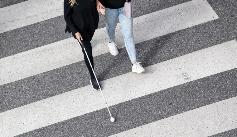 Scene of a Blind woman walking on zebra crossing helped by another person using her white cane. Help in the early stages of blindness