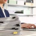 Stack of documents and woman working with laptop at table in office, closeup