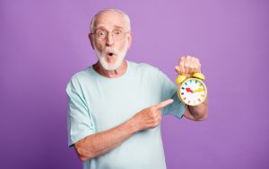 Portrait of funky amazed pensioner holding pointing in hand clock wear blue t-shirt isolated over purple color background