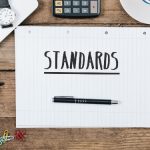 Headline Standards on note pad; office desk with electronic devices, computer and paper, wood table from above