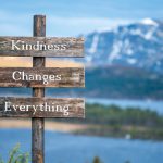 kindness changes everything text on wooden signpost outdoors in landscape scenery during blue hour. Sunset light, lake and snow capped mountains in the back.
