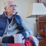 Older Man Wearing Extra Clothes With Hot Drink Trying To Keep Warm At Home In Energy Crisis