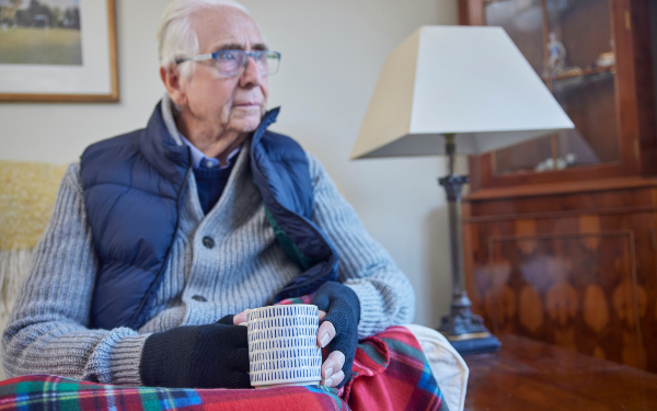 Older Man Wearing Extra Clothes With Hot Drink Trying To Keep Warm At Home In Energy Crisis