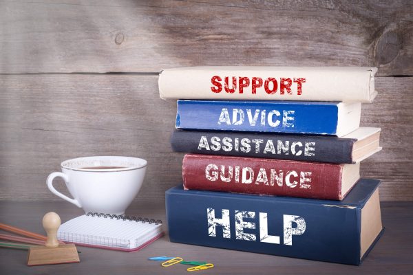 Pile of books on a wooden desk with the words 'advice', 'assistance', 'guidance' and 'help' on the spines