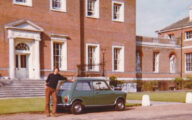 A tall young man, Tony Inwood, wearing brown trousers and a black top, leaning against a small green car, in front of a grand stately home