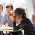 Indian woman laughing at funny joke eating pizza with diverse coworkers in office, friendly work team enjoying positive emotions and lunch together, happy colleagues staff group having fun at break