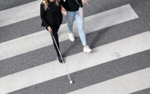 Scene of a Blind woman walking on zebra crossing helped by another person using her white cane. Help in the early stages of blindness