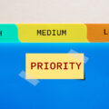 Folder with tabs headed 'high', 'medium' and 'low', above a post-it with the word 'priority' on it