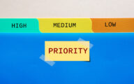 Folder with tabs headed 'high', 'medium' and 'low', above a post-it with the word 'priority' on it