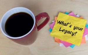 Mug full of coffee next to pile of post-it notes, with the top one reading 'What's your legacy?'