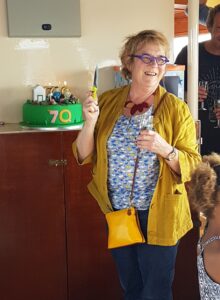 Pat Curtis wearing a yellow cardigan, blue top and jeans and smiling in front of a green cake that says '70'.