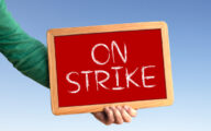Person's arm, with green sleeve, holding up red chalkboard with the word 'on strike' written in white chalk