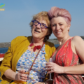 Pictured: (left) Pat Curtis smiling in a yellow cardigan and a blue dress with dots and Nicola Silk (right) in a pink dress with matching pink hair. Both are holding a drink and are seated on a boat in the sea.