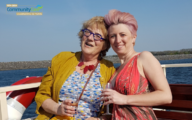 Pictured: (left) Pat Curtis smiling in a yellow cardigan and a blue dress with dots and Nicola Silk (right) in a pink dress with matching pink hair. Both are holding a drink and are seated on a boat in the sea.