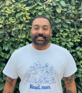 Khaleel, a team manager at Walsall. Khaleel is standing in front of a green leafy background, looking at the camera and smiling. He has short black hair and black stubble and is wearing a white t-shirt.