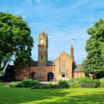 Walsall Arboretum. The image shows a striking building with a high clock tower set against a blue sky, with green grass at the front and two trees in full leaf either side.