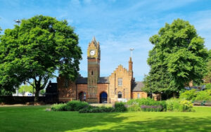 Walsall Arboretum. The image shows a striking building with a high clock tower set against a blue sky, with green grass at the front and two trees in full leaf either side.