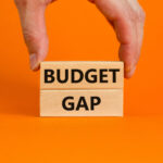 Person placing a wooden block with the word 'budget' written in black on it on top of a similar block with the word 'gap' written on it