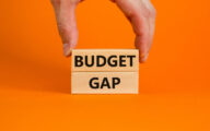 Person placing a wooden block with the word 'budget' written in black on it on top of a similar block with the word 'gap' written on it