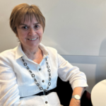 Pictured: Moira Gibb sitting against a white wall, wearing a white shirt with a black geometric necklace.