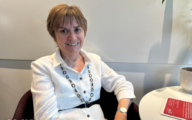 Pictured: Moira Gibb sitting against a white wall, wearing a white shirt with a black geometric necklace.