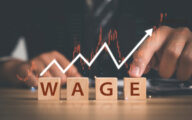 Blocks spelling out the word 'wage' with a line graph above it and a man in a suit pointing at it