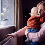 Mother With Son Trying To Keep Warm By Radiator At Home During Cost Of Living Energy Crisis