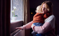 Mother With Son Trying To Keep Warm By Radiator At Home During Cost Of Living Energy Crisis