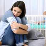 Young mother looking stressed in foreground with baby in cot in background