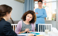 Social worker talking to, and going through paperwork with, parent/carer at a table, with son standing in background