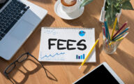 The word 'fees' written on a notepad in the middle of a desk, alongside a computer, phone and some pencils