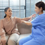 A care worker or nurse helping an older woman through an arm exercise, while both are sitting on a sofa in a living room