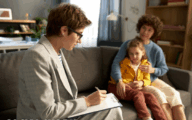 Social worker making notes in a family home with mother holding daughter on a sofa in the background