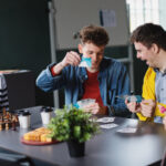 A couple of young men laughing while playing cards looked on by a woman playing chess and another woman knitting in a community centre